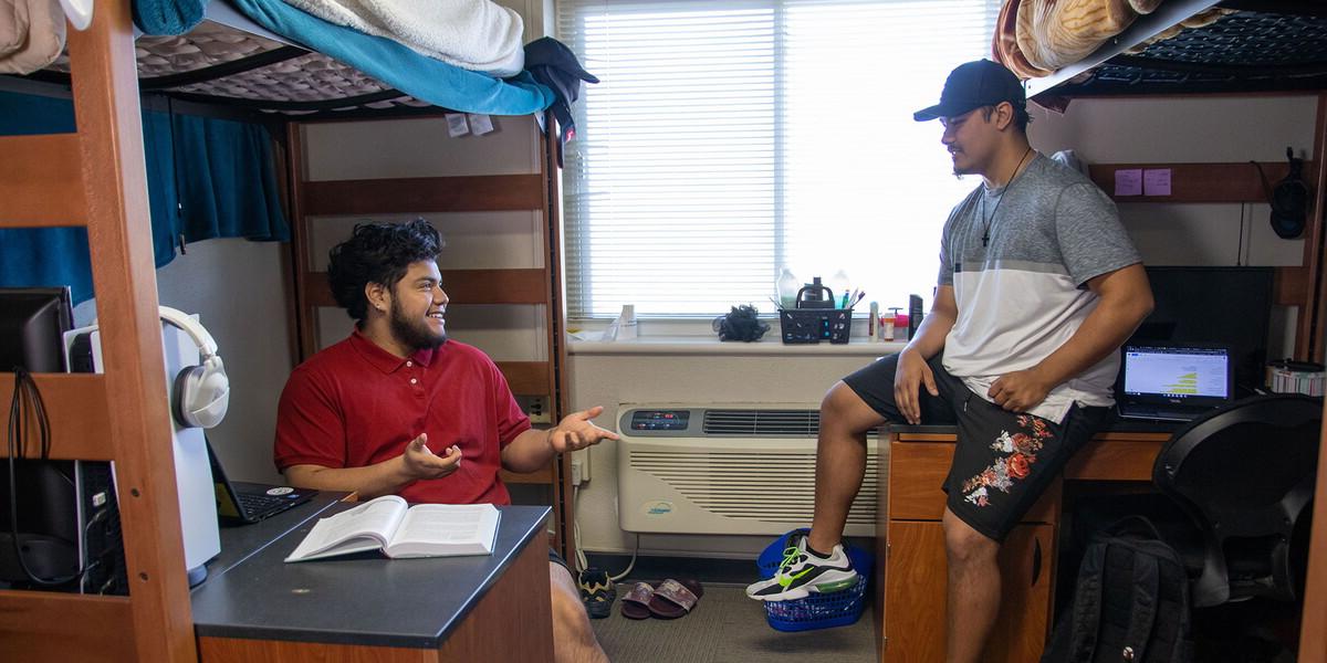 Two young men sitting on opposite sides of a dorm room under lofted beds.