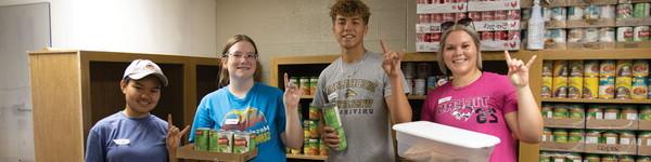 A group of students hold up pantry goods in front of shelves of food.